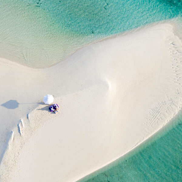 Aerial view of a sandbar surrounded by clear turquoise water at a Maldives luxury resort. Two people sit under a white umbrella on the sandbar's center, casting a shadow. The smooth white sand tapers off into the shallow waters around it, offering an idyllic setting reminiscent of Soneva's allure.