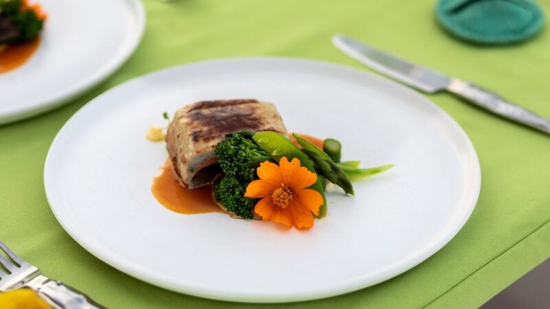 A neatly plated dish consisting of a rolled or wrapped main item, garnished with a bright orange flower, asparagus, snap peas, and broccoli atop a light sauce. The setting mirrors the elegance of a Maldives Luxury Yacht Holiday with its fork and knife on a lime green tablecloth.