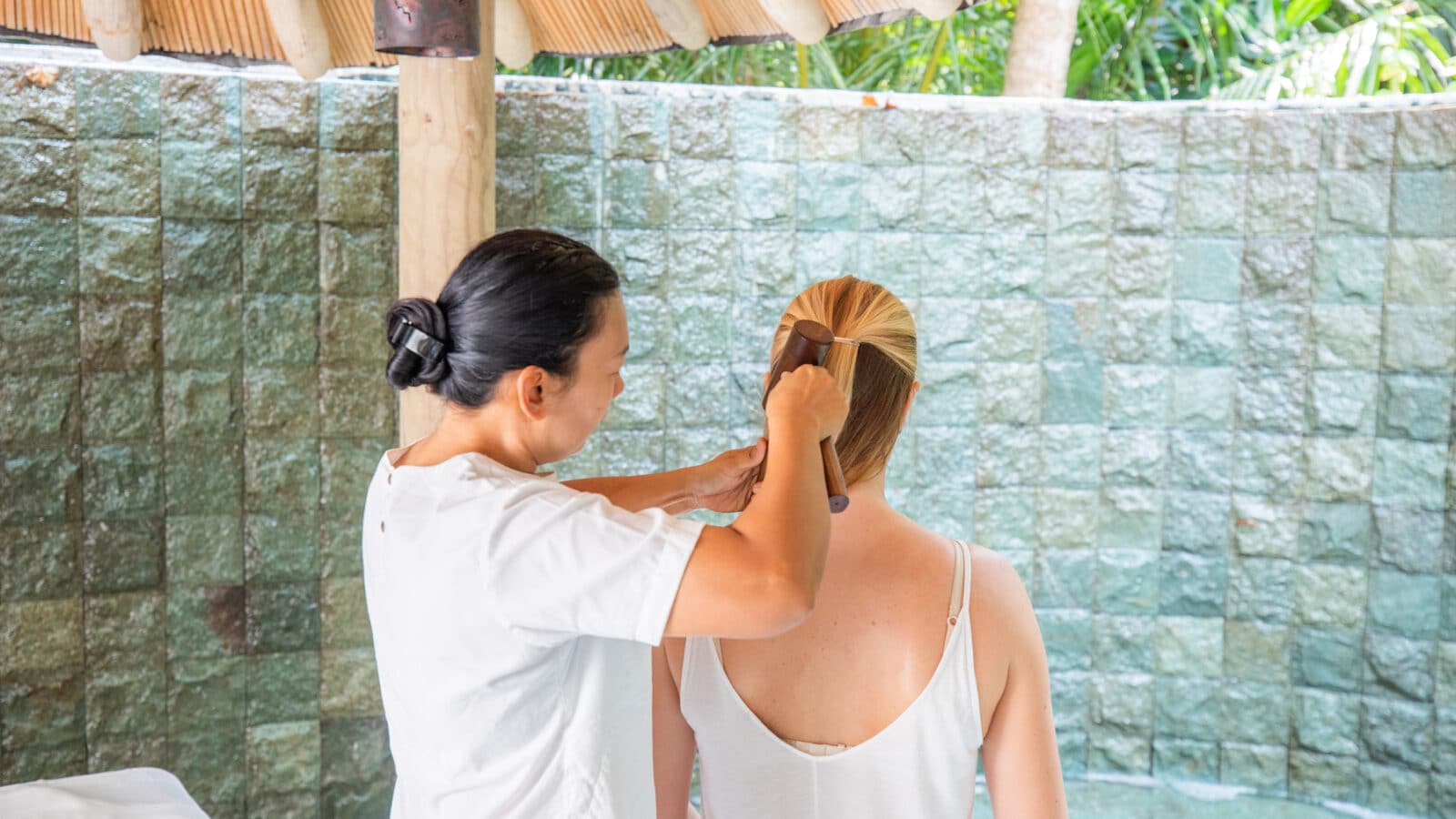 A woman in a white shirt is combing the hair of another woman who is sitting with her back facing the camera. They are at a serene Maldives spa, enveloped in an outdoor area with green tile walls and a thatched roof overhead, enjoying a moment of wellness.