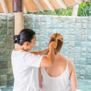 A woman in a white shirt is combing the hair of another woman who is sitting with her back facing the camera. They are at a serene Maldives spa, enveloped in an outdoor area with green tile walls and a thatched roof overhead, enjoying a moment of wellness.