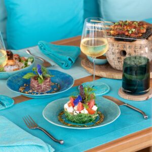 A turquoise-themed table setting features a vibrant seafood and vegetable dish, a glass of white wine, and a blue drink. The background includes more plates with various appetizers and a small wooden stove. Turquoise napkins and pillows complete the decor, reminiscent of Soneva Exclusive Offers.