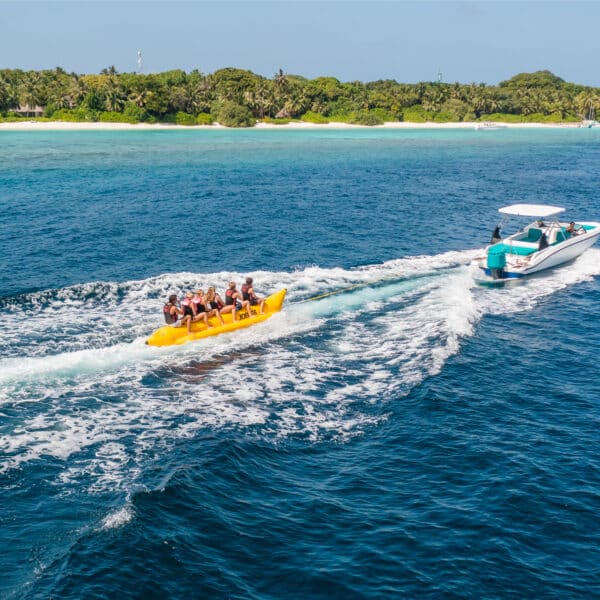 A group of people riding an inflatable banana boat is being pulled by a motorboat on clear blue ocean water. In the background, there is a lush green island with a sandy coastline and some scattered boats, part of the idyllic Soneva Exclusive Experience Maldives. The sky is clear and sunny.