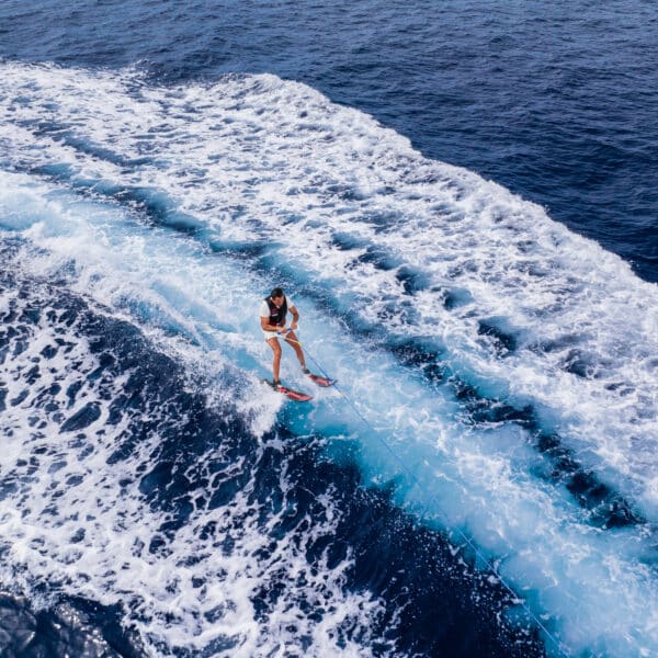 A person is riding a wakeboard on the ocean, being pulled forward and creating a trail of white water and foam. The deep blue water of this Soneva Exclusive Experience in the Maldives creates a stunning contrast with its foamy appearance, making the scene dynamic and refreshing.