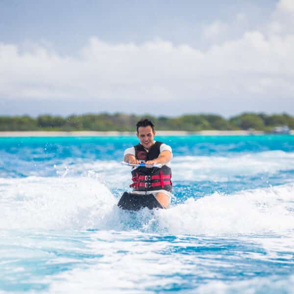 A person is wakeboarding on a bright, sunny day. They are wearing a red and black life vest and are being pulled rapidly across the water, creating splashes. In the background of this exclusive Soneva experience in the Maldives, there is a lush, green shoreline and a partly cloudy sky.