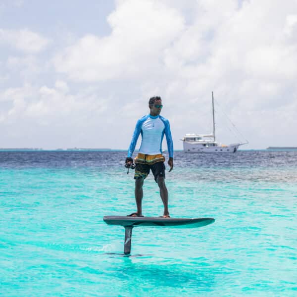 A person stands on an electric hydrofoil surfboard, gliding above turquoise water in the Maldives. Wearing sunglasses and a blue rash guard, the person is in a relaxed stance. A white sailboat is anchored in the distance under a partly cloudy sky, completing this exclusive Soneva experience.