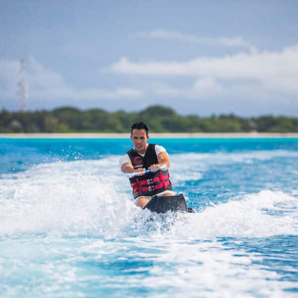 A person is enjoying a Soneva Exclusive Experience Maldives, riding a jet ski on clear blue water under a partly cloudy sky. They are wearing a life vest and appear to be relishing the sunny day, with a distant view of an island and some trees in the background.