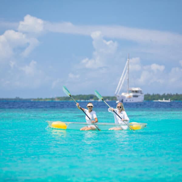 Two people are kayaking in a clear, turquoise sea under a bright blue sky with fluffy white clouds. Wearing sunhats and sunglasses, they paddle serenely as part of their Soneva Exclusive Experience Maldives. A sailboat is visible near the horizon, with a lush green island beyond the calm water.