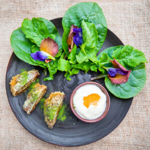 A beautifully arranged plate features fresh spinach leaves topped with edible flowers, a small dipping bowl filled with creamy white sauce, and a few pieces of fried vegetables covered in green seasoning. This exquisite dish, part of Soneva Exclusive Offers, rests on a textured burlap surface.