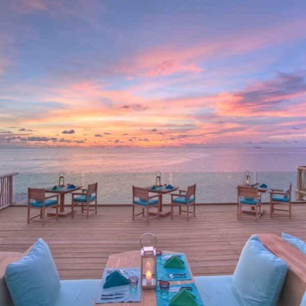 An outdoor dining area on a wooden deck at Soneva Fushi overlooks a calm ocean at sunset. Tables are set with blue chairs, napkins, and lanterns. The sky is painted with vibrant shades of pink, purple, and orange. A thatched roof structure is partially visible on the right.