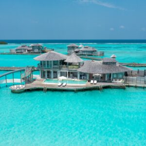 A luxurious overwater villa set on a turquoise ocean features multiple thatched-roof structures connected by wooden walkways. The villa includes a slide leading directly into the clear water and lounge chairs on the deck for relaxation, all part of the Soneva Exclusive Offers experience.