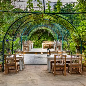 Outdoor dining area featuring wooden tables and chairs arranged under a metal archway covered with green foliage. Beyond the archway, a raised wooden deck with additional seating is visible, surrounded by lush greenery, creating a serene and inviting atmosphere reminiscent of Soneva Maldives luxury.