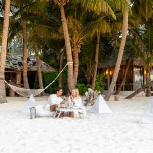 A man and woman sit at a small table on a sandy beach, surrounded by palm trees. They are enjoying drinks and are dressed in white. In the background, there is a rustic beach bar, a hammock, and several white pyramidal tents scattered around—an idyllic scene reminiscent of Soneva Exclusive Offers.