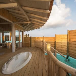 A luxurious overwater bungalow featuring a deck with a built-in bathtub, wooden railings, and views of the turquoise sea. The interior, part of Soneva Exclusive Offers, showcases cozy modern decor with large windows that let in abundant natural light.