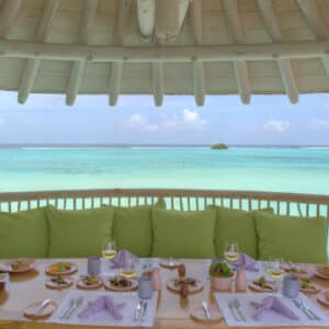 A dining table set for a meal under a wooden gazebo overlooking a serene turquoise ocean. The table is arranged with plates of food, glasses, baskets of bread, and napkin-wrapped cutlery. Green cushions line the seating area, contrasting with the clear blue sky and water—a scene worthy of Soneva Exclusive Offers.