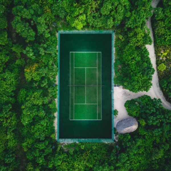 Aerial view of a secluded tennis court surrounded by dense green foliage and a small dirt path on the right. This Maldives exclusive experience features vibrant greenery contrasting with the dark green surface of the court, highlighting its rectangular shape and white lines.