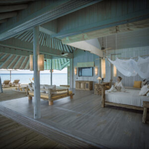 Open-air, rustic beachfront villa at Soneva in the Maldives with light blue wood beams and floor. A canopy bed is in the foreground, while seating areas are in the background, offering expansive views of the ocean. The decor is minimal and natural, enhancing the serene atmosphere of this luxury private resort.