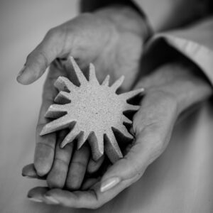 Black and white image of two hands cradling a textured, sun-shaped object with multiple pointed rays. The object looks handcrafted and is held gently, conveying a sense of care. Reminiscent of Soneva Exclusive Offers, the background is blurred, emphasizing the hands and the sun-shaped item.