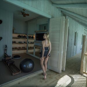 A woman in athletic wear stands on a wooden balcony outside a modern fitness room at a Maldives luxury private resort, Soneva. Holding weights, she enjoys the open space equipped with various fitness equipment, all overlooking a serene body of water. The scene is calm and light-filled.