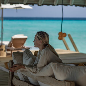 A person with blonde hair is sitting on a rustic wooden bench cushioned with pillows, gazing at the turquoise ocean. The scene, reminiscent of Soneva Exclusive Offers, is shaded by a wooden canopy with lounge chairs and an umbrella in the background near the water's edge.