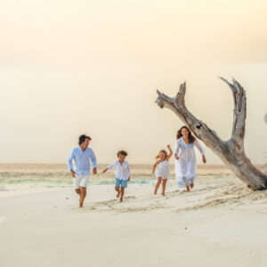 A family of four, dressed in white, joyfully runs along a sandy beach with the ocean in the background. The sky is overcast, and there is a large, weathered driftwood tree trunk on the right side of the image—capturing a perfect memory from their family holiday at Maldive Soneva.