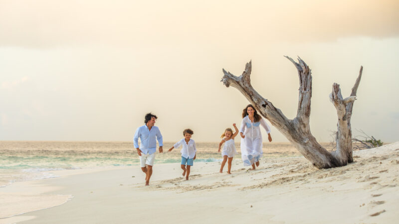 A family of four, dressed in white, joyfully runs along a sandy beach with the ocean in the background. The sky is overcast, and there is a large, weathered driftwood tree trunk on the right side of the image—capturing a perfect memory from their family holiday at Maldive Soneva.