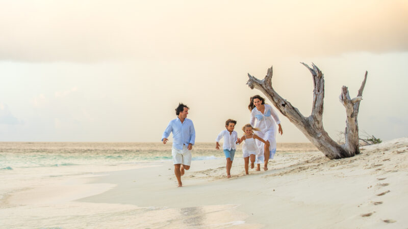 A family of four, dressed in light-colored clothes, is running along a sandy beach near the ocean during their Maldive Soneva holiday. The parents and two children appear joyful as they enjoy their time together. A weathered, leafless tree stands on the beach near them under a pastel-colored sky.