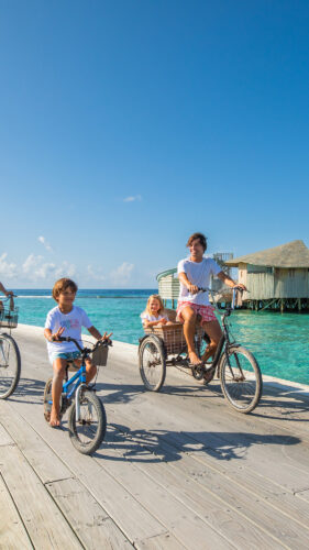 A family of four rides bicycles on a wooden boardwalk over clear turquoise water, likely in a tropical location. Two adults and two children, one in a child seat, smile and enjoy the sunny weather. Water villas are visible in the background, making this the perfect family holiday at Soneva in the Maldives.