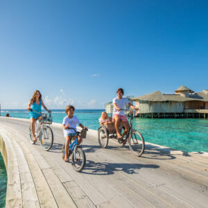 A family of four rides bicycles on a wooden boardwalk over clear turquoise water, likely in a tropical location. Two adults and two children, one in a child seat, smile and enjoy the sunny weather. Water villas are visible in the background, making this the perfect family holiday at Soneva in the Maldives.