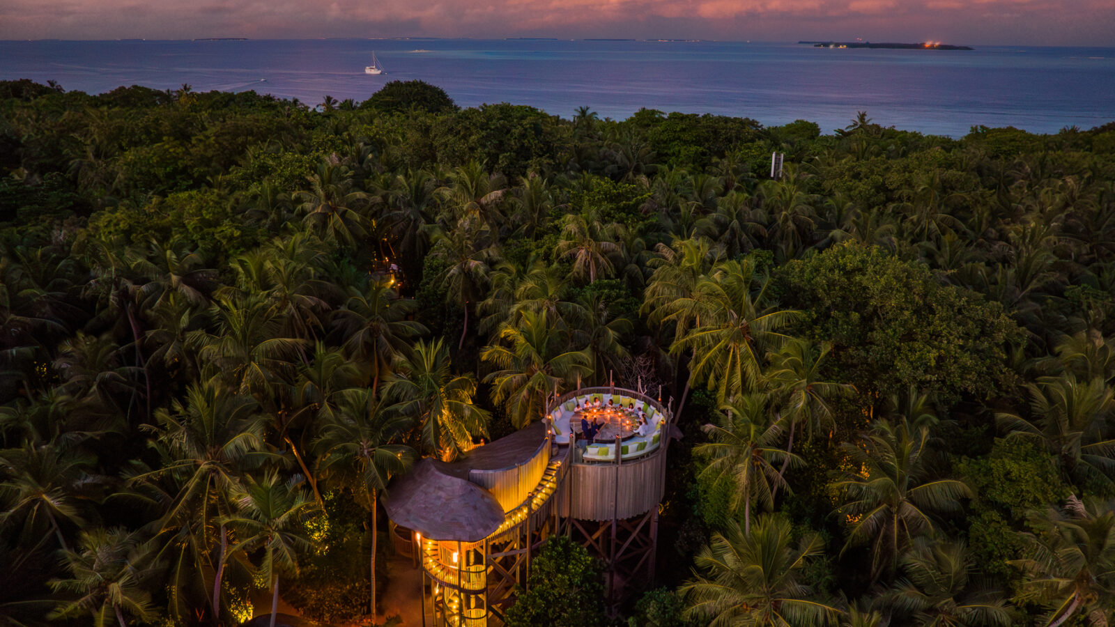 Aerial view of a treehouse restaurant nestled among dense palm trees at sunset. The elevated dining area glows warmly with lights, overlooking a vast forest landscape and the calm ocean beyond, beneath a twilight sky with subtle orange and pink hues—true Maldives dining at Soneva Fushi.