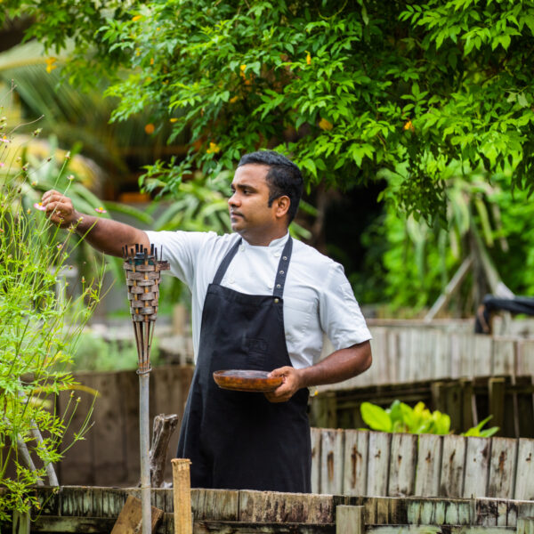 A man in a white shirt and black apron stands in a lush garden at Soneva Fushi, holding a wooden bowl and reaching out to touch tall plants. A tiki torch and wooden fence are visible nearby, with green trees and foliage filling the background, evoking the essence of Maldives dining amidst nature.