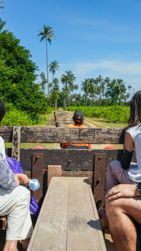 Five people are sitting on a wooden open-air cart being pulled by a tractor through a tropical landscape with lush greenery and palm trees under a clear blue sky, enjoying an exclusive Thailand experience offered by Soneva.