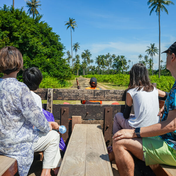 Five people are sitting on a wooden open-air cart being pulled by a tractor through a tropical landscape with lush greenery and palm trees under a clear blue sky, enjoying an exclusive Thailand experience offered by Soneva.