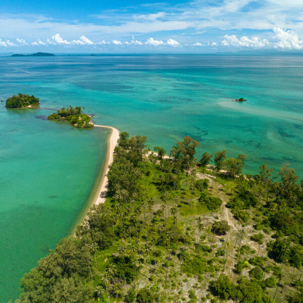 Aerial view of lush, green islands surrounded by clear turquoise waters. The shoreline features sandy beaches and dense palm trees. The sky is clear with a few scattered clouds, and the tranquil sea stretches into the horizon, offering a Thailand exclusive experience at Soneva resorts.