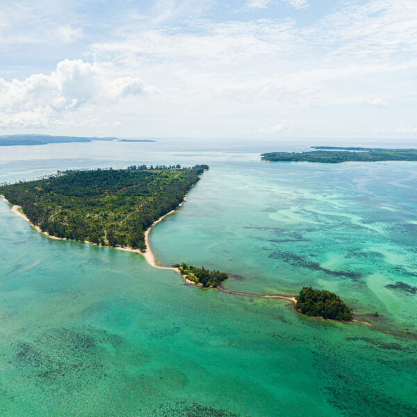 Aerial view of a tropical island in Thailand surrounded by turquoise waters and coral reefs. The island, part of the exclusive Soneva experience, is lush with vegetation, featuring sandy shores and a smaller islet connected by a narrow strip of land. The horizon reveals more islands and a partly cloudy sky.