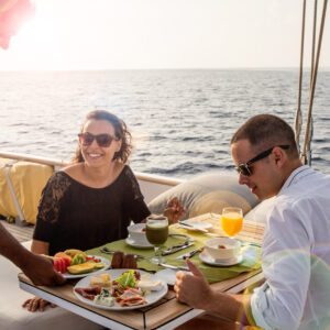 A couple enjoys a meal on a luxury yacht at Soneva in the Maldives, with a server presenting their food. The woman, wearing sunglasses, smiles at the server. The man, also in sunglasses, looks at his plate. The table is set with various dishes and drinks as the ocean and sunlight create a perfect backdrop.