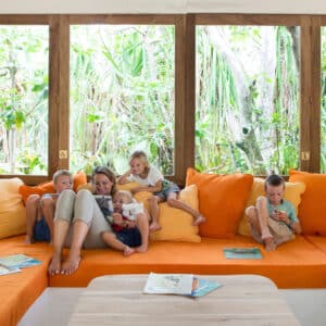 A woman and five children sit on an orange cushioned bench in front of large windows displaying lush greenery. Books and toys are scattered around. The woman and four children engage with a baby, while one child is distracted on the side. It&#039;s a serene moment reminiscent of Soneva Exclusive Offers.