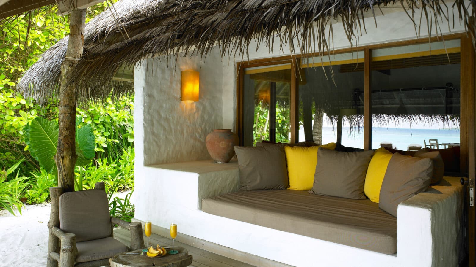 A cozy outdoor seating area at the Maldives Luxury Resort features a cushioned bench with yellow and gray pillows under a thatched roof. Nearby, there's a rustic wooden chair and table with two glasses of orange juice. Lush greenery surrounds the space, and soft ambient light illuminates the scene.