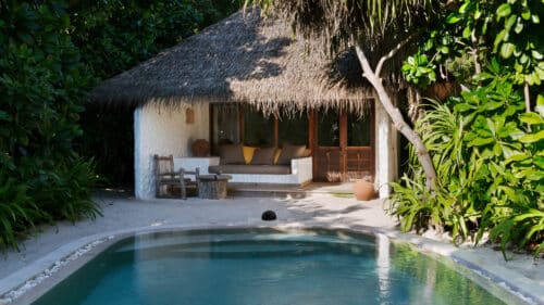 A cozy bungalow with a thatched roof is nestled among lush green plants at the Maldives Luxury Resort, Soneva Fushi Villas. The bungalow features a shaded seating area with cushioned furniture and wooden chairs. A serene outdoor swimming pool is situated in the foreground, surrounded by a sandy area.