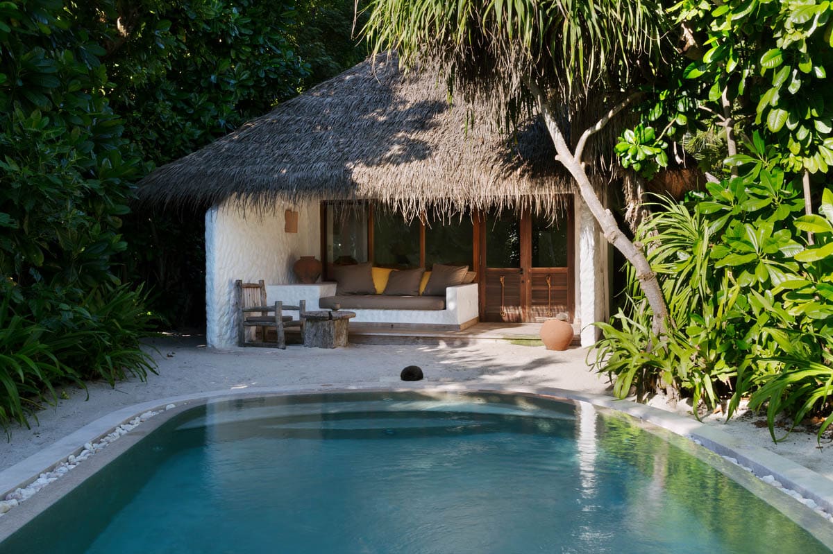 A cozy bungalow with a thatched roof is nestled among lush green plants at the Maldives Luxury Resort, Soneva Fushi Villas. The bungalow features a shaded seating area with cushioned furniture and wooden chairs. A serene outdoor swimming pool is situated in the foreground, surrounded by a sandy area.