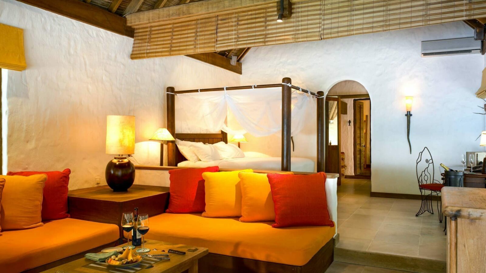 A cozy, rustic bedroom reminiscent of a Maldives luxury resort with wood-beamed ceilings. The room features a four-poster bed draped with a white canopy, a wooden table with wine and glasses, vibrant cushioned couches, and warm ambient lighting. The walls are white and there's a wooden door.