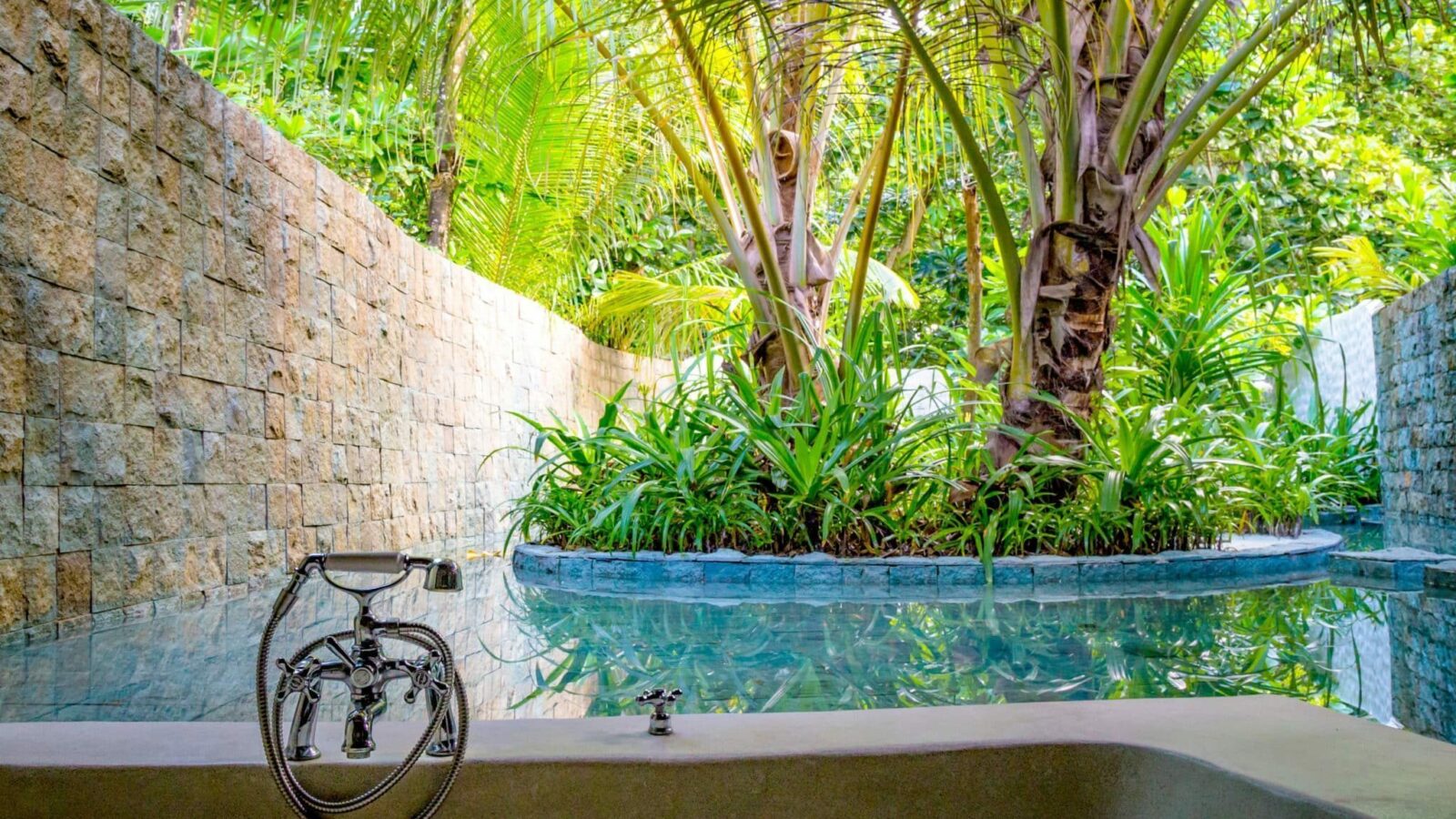 A serene outdoor bathtub is set in a lush, tropical garden at Maldives' luxury resort, Soneva Fushi Villas. The stone walls enclose the space, while palm trees and various plants surround a clear pool of water. The bathtub faucet is prominently featured in the foreground.