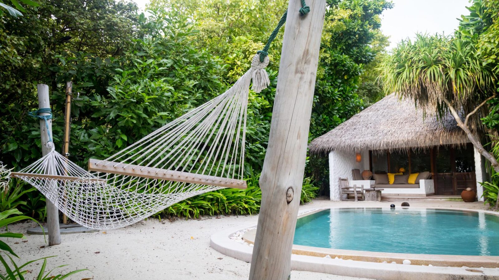 A serene outdoor scene features a hammock tied between two wooden poles on a sandy area next to a turquoise swimming pool. In the background, there's a cozy thatched-roof hut surrounded by lush green foliage, reminiscent of the tropical, relaxing atmosphere found at Maldives luxury resorts like Soneva Fushi Villas.