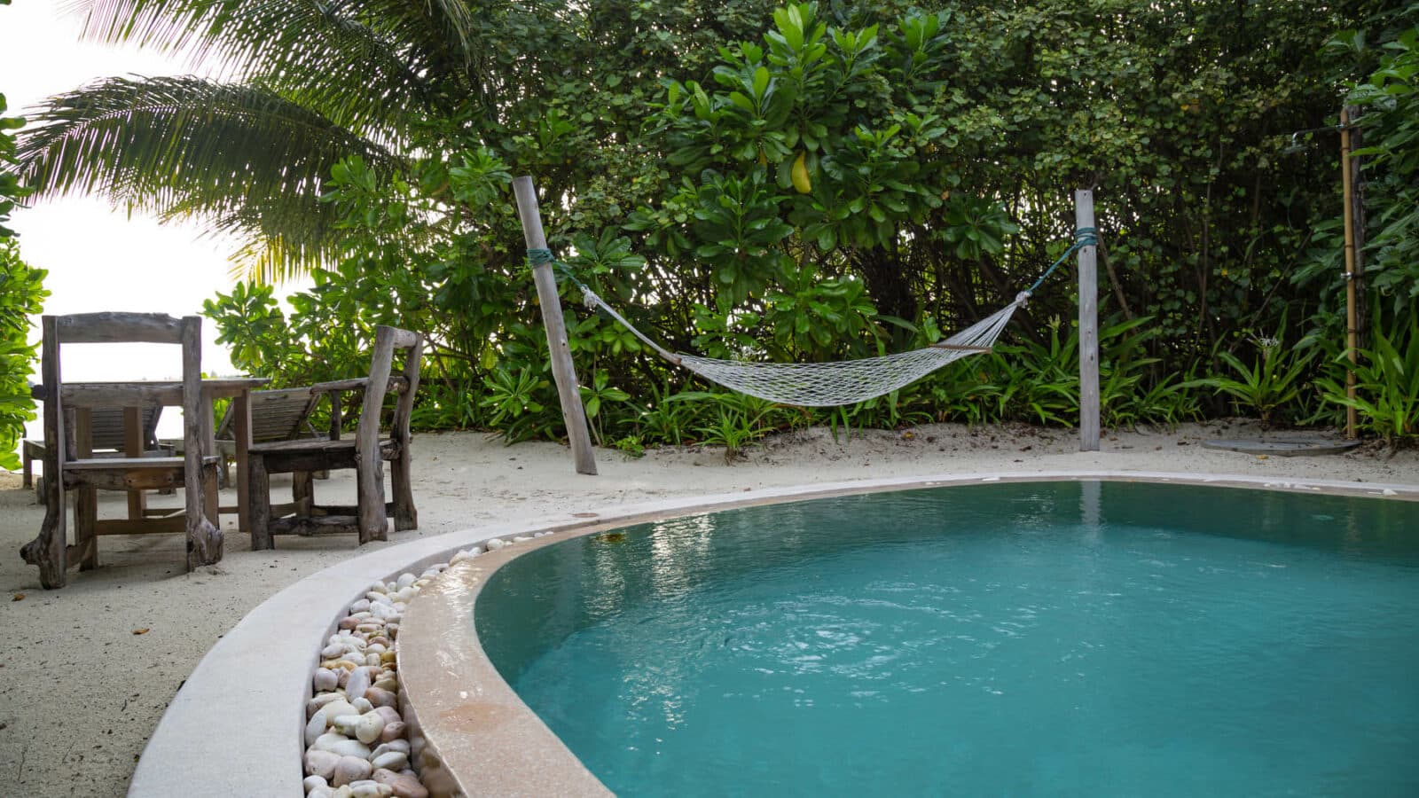 A serene tropical setting featuring a turquoise swimming pool with a stone border, adjacent to a hammock strung between two posts, and a rustic wooden table and chairs. Lush green foliage and palm trees surround the area, reminiscent of the exquisite Soneva Fushi Villas in a Maldives luxury resort.