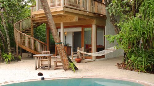 Experience a Maldives luxury resort at its finest with this tropical outdoor scene featuring a two-story wooden hut complete with a spiral staircase. The ground level boasts a patio adorned with wooden furniture and a hammock. Enveloped in greenery, the circular pool lies amidst the sand, reminiscent of Soneva Fushi Villas.