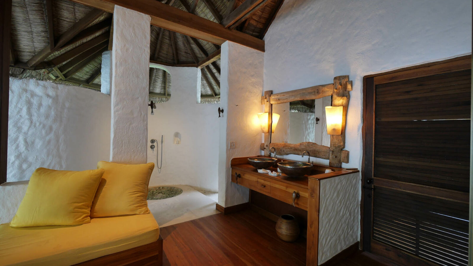 A cozy, rustic bathroom at the Maldives Luxury Resort features a high thatched ceiling. The room in Soneva Fushi Villas includes a wooden vanity with two sinks, warm lighting, a yellow cushioned seating area, and a stone-tiled open shower in the corner. White walls contrast elegantly with wood and natural tones.