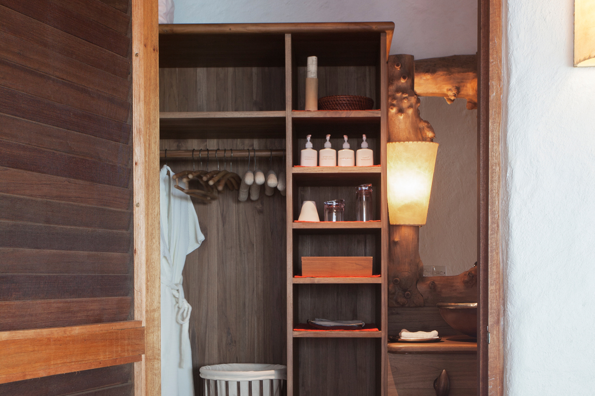 A cozy bathroom with rustic wooden shelves stocked with various bottles, towels, and toiletries evokes the charm of a Maldives luxury resort. A white robe hangs on a wooden hanger in a closet. A basket sits on the floor. There is a wooden-framed mirror and a softly glowing wall light, adding warmth to the space akin to Soneva Fushi Villas.