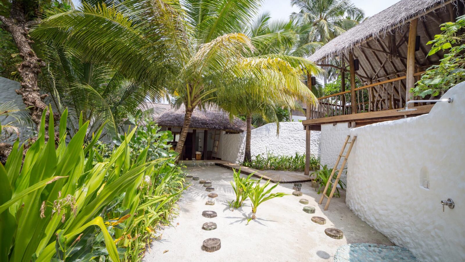 A tropical outdoor area with palm trees, lush green plants, and a sandy pathway lined with circular stepping stones leading to a thatched-roof hut. A wooden ladder leans against the side of another hut at the Maldives luxury resort, Soneva Fushi Villas, exuding a serene and natural ambiance.