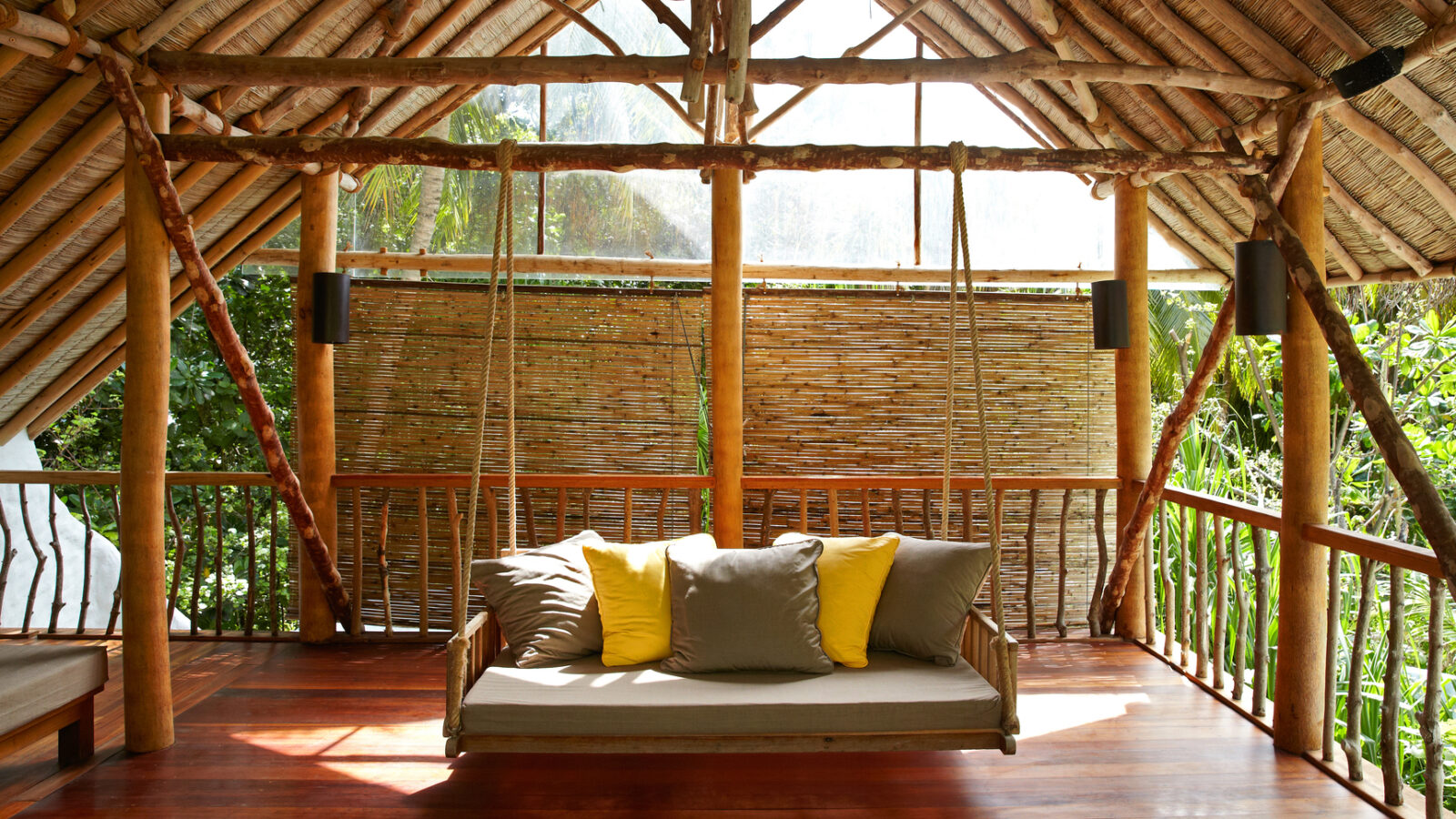 An open-air, rustic wooden loft with a thatched roof, featuring a suspended wooden swing-bed adorned with gray and yellow pillows. Bamboo shades and lush greenery are visible through the gaps, creating a serene, nature-inspired space reminiscent of the Maldives' Soneva Fushi Villas.