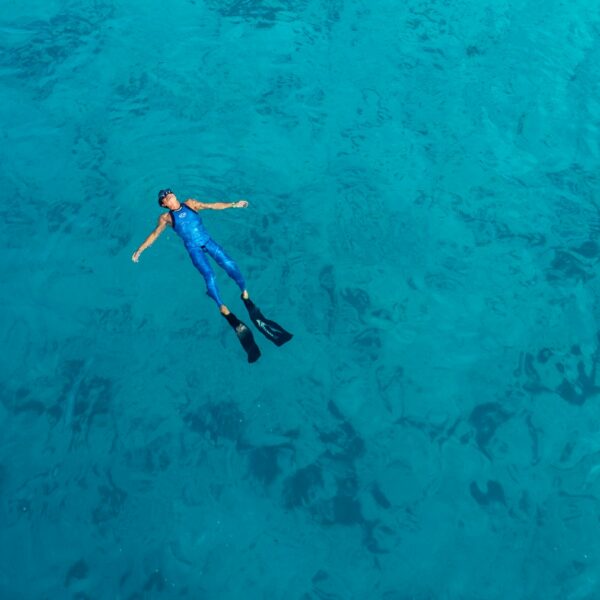 A person wearing a blue wetsuit and black flippers floats on their back in clear, blue water during their Maldives luxury yacht holiday. They appear relaxed, with arms outstretched and legs extended in the vast expanse of the serene sea.