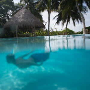 A person swims underwater in a clear, blue pool surrounded by tropical palm trees. The luxurious Soneva Fushi Villas are visible in the background, with their thatched roofs, lush greenery, and bright sky contributing to the serene Maldives vacation atmosphere.